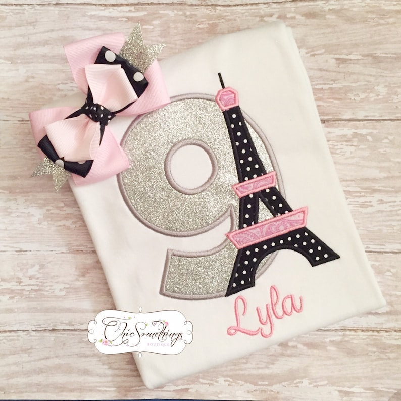 May include: A white t-shirt with a silver glitter number 9 and a black and pink Eiffel Tower design. The name "Lyla" is embroidered below the design. A pink and black polka dot bow is on the shirt.