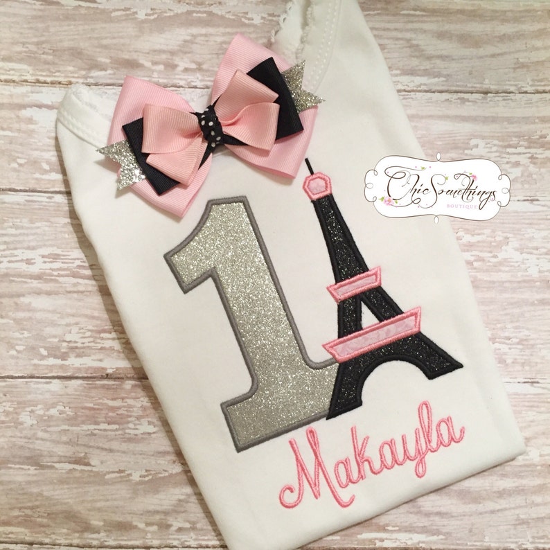 May include: A white onesie with a silver glitter number one, a pink and black bow, and a black and pink Eiffel Tower design. The onesie is personalized with the name "Makayla" in pink.