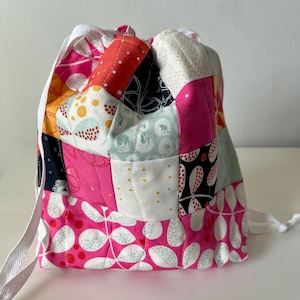 May include: A patchwork drawstring bag with a variety of colorful fabric squares. The bag features pink, red, white, and navy blue patterns, with a white drawstring closure and straps. The bag is sitting on a white surface.