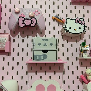 Könnte beinhalten: Eine hellrosa Lochwand als Wandorganizer mit einem pastellrosa Gaming-Controller mit herzförmiger Schleife, einem Hello Kitty-Wandbecher für Häkelnadeln, einer zweischübigen Aufbewahrungsbox im Katzen-Design und einem Regal mit Pfotenabdruck-Motiv.