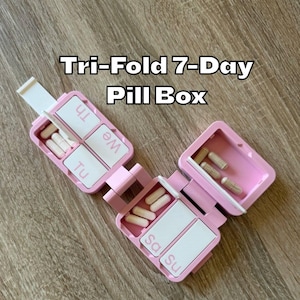 Könnte beinhalten: Eine rosa, dreifach gefaltete 7-Tage-Pillenbox, geöffnet, um Fächer für jeden Wochentag anzuzeigen. Die Box ist mit dem weißen Text "Tri-Fold 7-Day Pill Box" beschriftet. Die Fächer enthalten Pillen.