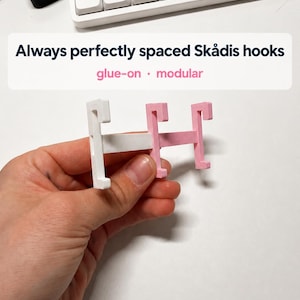 Op de afbeelding: Een witte en lichtroze modulaire haakverbinder met de tekst Always perfectly spaced Skådis hooks glue-on modular. Het object heeft twee verticale haakcomponenten verbonden door een horizontale balk, ontworpen voor opbergsystemen.