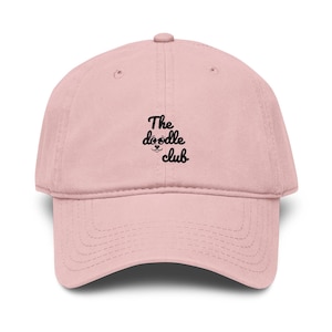 May include: A light pink baseball cap featuring a structured crown and a curved brim. Centered on the front panel is black embroidered text that reads The doodle club, accompanied by a small graphic of a dog nose and mouth below the word doodle.