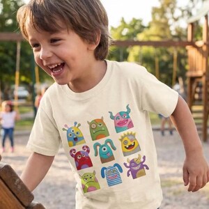 May include: A cream-coloured short-sleeve t-shirt featuring a graphic print of nine distinct colourful cartoon monsters. The monsters are arranged in a grid pattern and display various expressions, shapes, and patterns in shades of blue, green, red, and purple.
