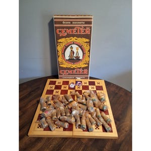 May include: A wooden chessboard with a red and light wood checkered pattern, featuring small cross symbols in each square. A tall, rectangular box with the text "СЕМЕТЕЙ" and "белек шахматы" stands behind the board, containing wrapped chess pieces.