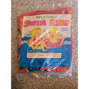 Könnte beinhalten: Ein aufblasbarer Schwimmring in einer durchsichtigen Plastikverpackung. Das Etikett enthält den Text INFLATABLE SWIM RING, SIZE: 20" APPROX und CAUTION: AGE 3 UP. Die Grafik zeigt zwei Kinder in Schwimmringen mit Wasser-, Blumen- und Krabbenmotiven.