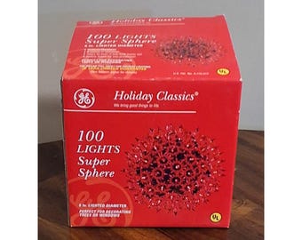 May include: A red GE Holiday Classics product box featuring a 100 light super sphere with a 6 inch lighted diameter. The packaging displays the GE logo, text reading 100 Lights Super Sphere, and a graphic of a spherical light fixture.
