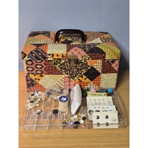 Puede incluir: Una caja de costura rectangular con cubierta de tela de patchwork en tonos naranja, marrón y amarillo, con asa marrón oscuro y cierre metálico. Debajo, una bandeja organizadora de plástico transparente contiene botones y carretes.