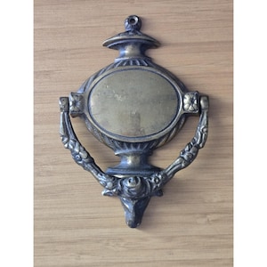 May include: An antique brass door knocker featuring an oval central plate with a smooth finish. The frame is decorated with floral motifs and scrollwork, including a prominent rose detail at the base. It has a mounting hole at the top.