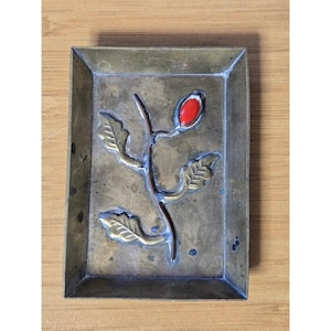 May include: A rectangular brass tray featuring a raised relief design of a single stem with three leaves and a prominent, smooth, bright red berry. The metal surface shows a weathered, aged patina with dark spots and a matte, metallic finish.
