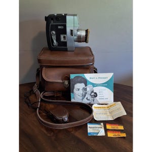 May include: A vintage Bell & Howell Optronic Eye 8mm movie camera with a brown leather carrying case, an instruction manual, a small brown leather accessory case, and several pieces of film packaging including Dynachrome and Kodachrome labels.