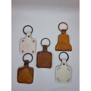 May include: Five leather keychains with metal rings. Two are white with floral accents and letters R and K. Two are brown with floral accents and letters A and J. One is a brown bell-shaped keychain with the text Let Freedom Ring embossed.
