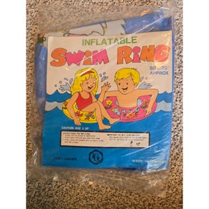 Może przedstawiać: Dmuchane koło do pływania zapakowane w przezroczystą folię. Na opakowaniu znajduje się napis INFLATABLE SWIM RING, SIZE 20 APPROX oraz ilustracja dwojga dzieci z kołami do pływania. Produkt wyprodukowany na Tajwanie, numer artykułu 10604PB.