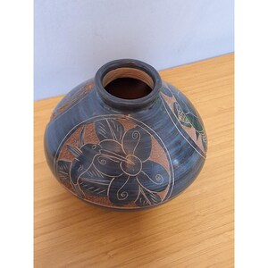 May include: A round, dark blue ceramic vase with a wide opening and a short neck. The surface features etched, light brown circular panels containing detailed carvings of a hummingbird and a large flower with swirling petal designs.