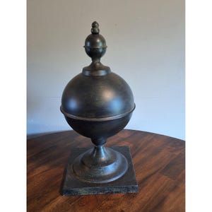 May include: A decorative finial object featuring a large central sphere resting on a pedestal base. The item is finished in a dark, distressed black and bronze metallic tone, topped with a smaller spherical accent and a pointed finial tip.