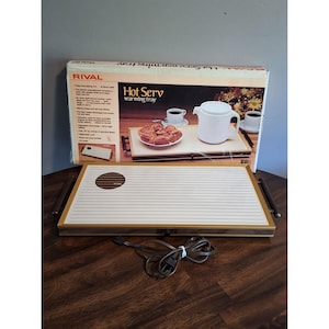 Puede incluir: Una bandeja calentadora rectangular Rival Hot Serv con superficie blanca de líneas horizontales, marco dorado y asas marrón oscuro a los lados. Incluye un logotipo circular marrón a la izquierda y un cable eléctrico de color negro.