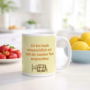 Op de afbeelding: Een witte keramische mok met een lichtgele achtergrond voorzien van de Duitse tekst "Ich bin heute voraussichtlich erst nach der zweiten Tasse ansprechbar" en een eenvoudige lijntekening van een persoon die in bed slaapt onder een geruite deken.