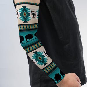 May include: An arm sleeve featuring a repeating pattern of teal and cream geometric shapes, black buffalo silhouettes, and bull skull icons. The design includes horizontal bands of orange and teal diamonds against a dark teal background.