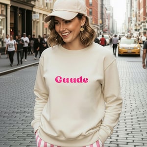 Könnte beinhalten: Ein cremefarbenes Sweatshirt mit dem Wort "Guude" in leuchtend rosa Buchstaben. Die Person trägt eine beige Baseballkappe und eine Hose mit rosa und weißen Streifen. Der Hintergrund ist eine Stadtstraße mit Menschen und Gebäuden.