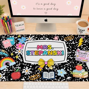 May include: A rectangular desk mat featuring a black and white composition notebook pattern with colorful illustrations of a rainbow, apples, books, a yellow bow, and a central white label reading MRS. STEPANSKI in multi-colored block letters.
