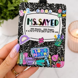 May include: A rectangular ID badge holder with a black and white composition notebook pattern. The centre features the text MS. SAYED, 100 Sheets, 200 Pages, 9 3/4 in x 7 1/2 in (24.7 cm x 19 cm), and colourful school-themed stickers including a calculator, scissors, and the text I LOVE MATH.