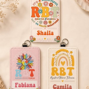 Op de afbeelding: Drie rechthoekige badgehouders met metalen ringen. De bovenste houder toont de tekst 'IT'S A BEAUTIFUL DAY TO SHAPE BEHAVIORS' met 'RBT' en 'Shaila'. Linksonder een zonnebloem met puzzelstukjes, 'RBT' en 'Fabiana'. Rechtsonder een regenboog, 'RBT' en 'Camila'.