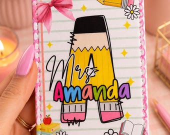 Könnte beinhalten: Ein personalisierter Lehrer-Ausweishalter mit Bleistift-Design. Der Ausweishalter zeigt den Namen "Mrs. Amanda" in bunten Buchstaben, eine große gelbe Bleistiftgrafik und dekorative Elemente wie eine Schleife, Blumen und Bücher. Der Ausweishalter ist rechteckig mit einem rosa genähten Rand.