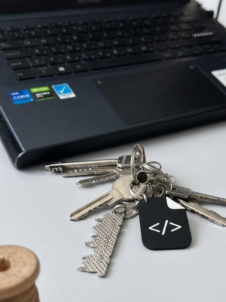 May include: A metal keyring featuring a black rectangular tag with a white code symbol, a silver textured skyline charm, and several silver keys. The keyring is positioned in front of a dark laptop computer with a visible keyboard and trackpad.