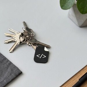 May include: A metal keyring featuring a black, rectangular tag with a white code symbol consisting of a slash between two angle brackets. The tag has a small white corner detail and is attached to a silver metal ring holding several silver keys.