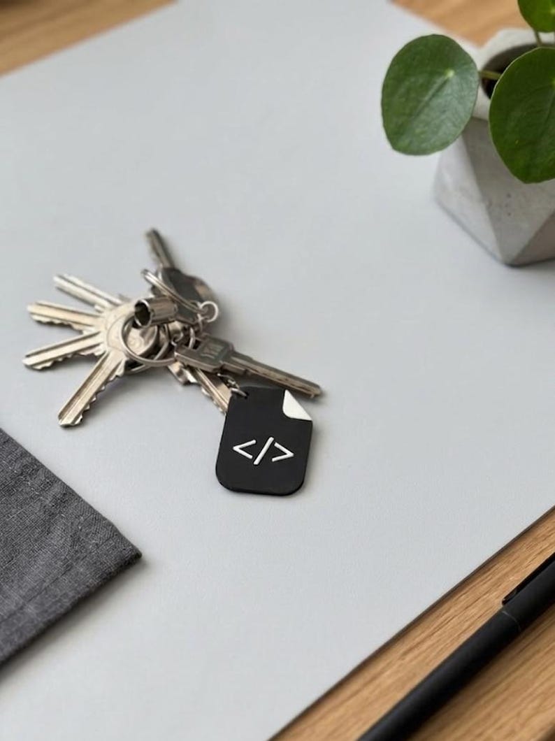 May include: A metal keyring featuring a black, rectangular tag with a white code symbol consisting of a slash between two angle brackets. The tag has a small white corner detail and is attached to a silver metal ring holding several silver keys.