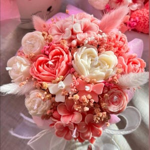 Könnte beinhalten: Ein Bouquet aus handgefertigten Blumenkerzen in Rosa-, Pfirsich- und Weißtönen. Das Arrangement umfasst rosenförmige Kerzen, kleine Blumenakzente und dekorative Schleifen, alles in rosa Papier verpackt und mit einem weißen Band gebunden.