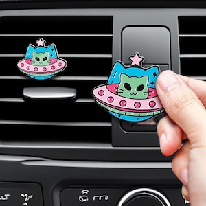 May include: Two car air freshener clips featuring a light green cat head inside a blue and pink UFO saucer. The cat has black eyes and whiskers, and a small pink star sits atop the blue cat ears. The design is colorful with a black outline.
