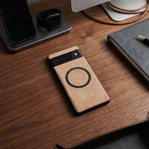 Può includere: Una custodia per telefono beige con una barra della fotocamera nera e un cerchio nero con testo. La custodia è su una superficie di legno, accanto a un orologio nero, un telefono nero e un taccuino grigio.