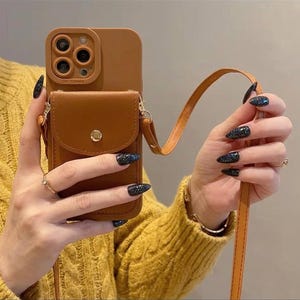 May include: A brown phone case with a small pouch and a long strap. The case has a camera cutout and a gold button closure on the pouch. The strap is tan and the case is held by a person wearing a yellow jumper.