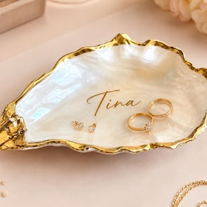 May include: A white, iridescent oyster shell trinket dish with a gold-painted, scalloped edge. The interior surface features the name Tina written in gold script. Two gold rings and a pair of small gold stud earrings rest inside the shell dish.