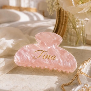 May include: A light pink hair claw clip with a marbled, pearlescent finish. The clip features the name Tina written in elegant, gold-colored cursive script across the center. The accessory has a curved, semi-circular shape with interlocking teeth.