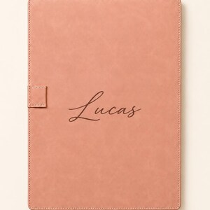 May include: A rectangular, dusty rose-coloured notebook with a smooth, matte finish. The cover features the name Lucas written in a dark brown, cursive script font. A small, matching rectangular pen loop is attached to the left side of the cover.