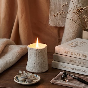 Puede incluir: Una vela de cerámica moteada y encendida con una superficie texturizada sobre una mesa de madera. Un plato pequeño con piedras y vegetación, y libros apilados con los títulos "Art & Nature", "Terres Cuites" y "L'Harmonie du Foyer" también están presentes.