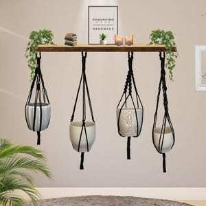 Puede incluir: Una estantería de madera con cuatro macetas colgantes suspendidas por macramé negro. Las macetas contienen varias plantas en maceta. La estantería también muestra pequeños libros, velas y una impresión enmarcada que dice "SimplyKorded Handmade."