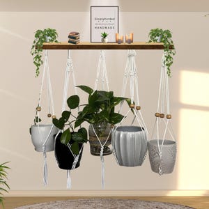 Puede incluir: Un colgador de plantas de macramé con cinco macetas de cerámica, cada una con una planta diferente. Las macetas son blancas, negras, grises y de vidrio transparente. Una estantería de madera encima contiene pequeñas plantas, velas y un letrero enmarcado "SimplyKorded HANDMADE."