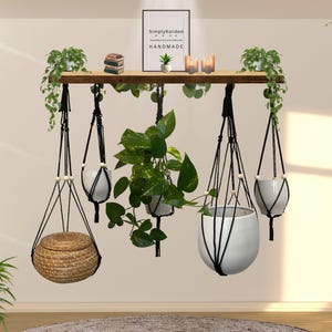 Puede incluir: Una estantería de madera con macetas colgantes de macramé, cada una con una maceta de cerámica blanca o una cesta tejida. La estantería también muestra pequeñas plantas, velas y una impresión enmarcada que dice "Simply Korded HANDMADE."