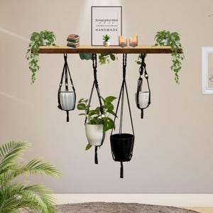 May include: A wooden shelf with hanging macrame planters, each holding a plant. The shelf also displays candles, a small plant, and a framed print that reads "SimplyKorded HANDMADE". The wall is a neutral colour, and there is a framed image of a person meditating.
