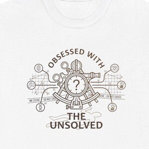 May include: A white t-shirt featuring a brown graphic of a sextant with a question mark in the center. The text reads "OBSESSED WITH THE UNSOLVED" and includes icons of handcuffs, a fingerprint, a camera, a handprint, and a DNA strand.