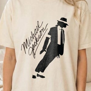May include: Cream-colored t-shirt featuring a black silhouette of Michael Jackson in a signature pose, with the name "Michael Jackson" in cursive script. The shirt is a casual, oversized fit.