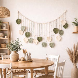 May include: A macrame leaf wall hanging in shades of green and cream, suspended from a wooden dowel. The leaves are arranged in a curved pattern, adding a touch of bohemian decor to a dining room setting. A wooden table and chairs complete the scene.