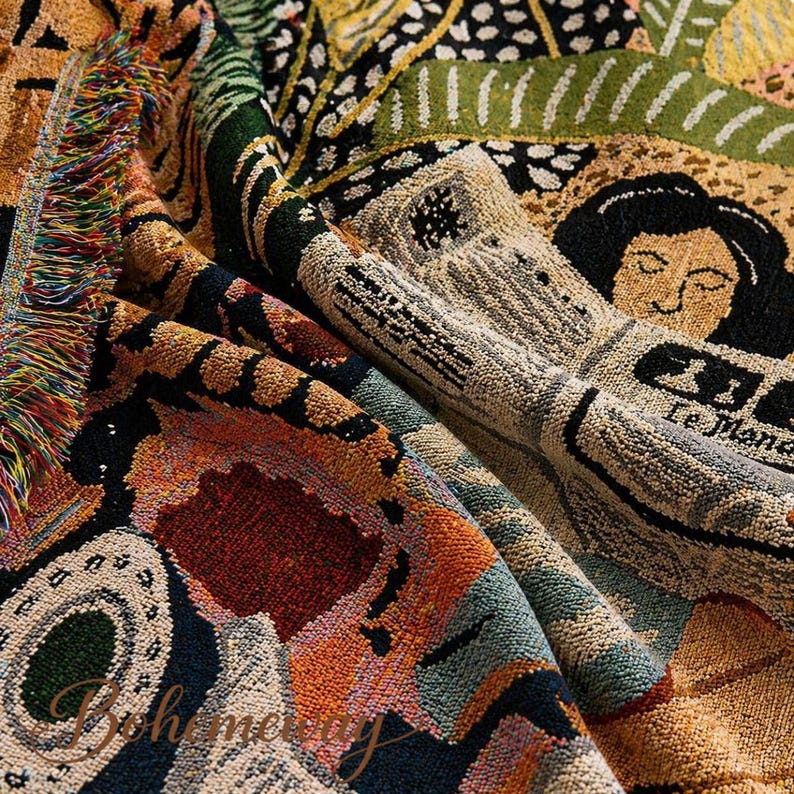 May include: A colorful woven throw blanket with a detailed pattern. The design incorporates various shapes, figures, and text in shades of orange, green, black, and beige. The blanket has a fringed edge and a textured appearance, suitable for home decor.