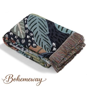 May include: Folded woven throw blanket with a botanical pattern in shades of blue, green, and black. The blanket has a fringed edge and the word "Bohemeway" in a script font.