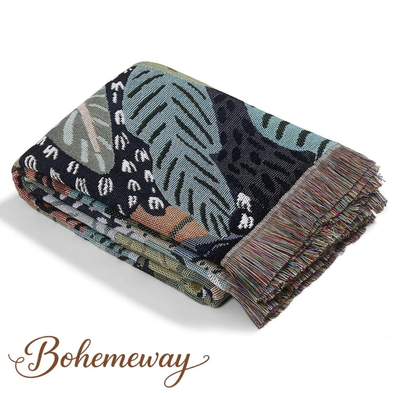 May include: Folded woven throw blanket with a botanical pattern in shades of blue, green, and black. The blanket has a fringed edge and the word "Bohemeway" in a script font.