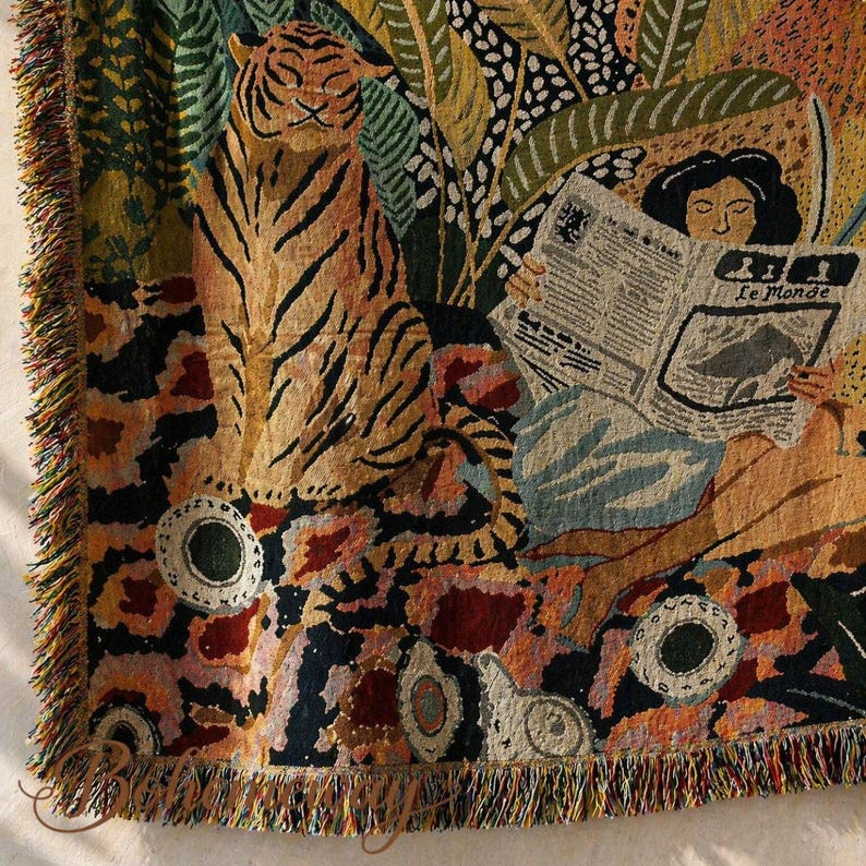 May include: Woven throw blanket featuring a tiger, a person reading "Le Monde", and jungle foliage. The blanket has a fringed edge and a colorful, patterned design with shades of orange, green, and blue. Decorative home decor.