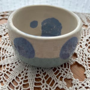 Könnte beinhalten: Kleine, handgefertigte Keramikschale mit cremefarbenem Innen- und Außenbereich. Die Schale hat große, hellblaue Tupfen auf der Außenseite und ein hellblaues Band am Boden. Wahrscheinlich zum Servieren kleiner Portionen von Speisen.